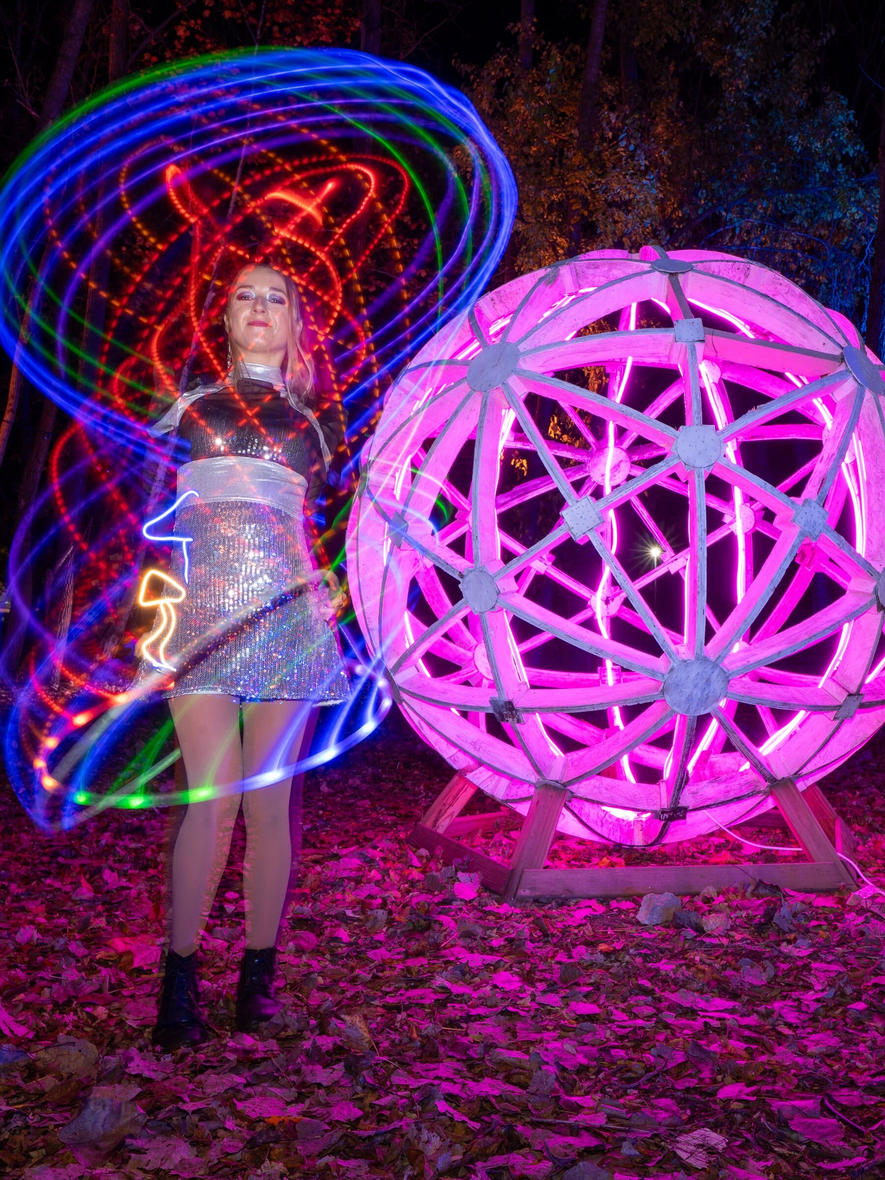 A woman spinning light-painting rings beside a glowing pink geodesic sphere in the woods
