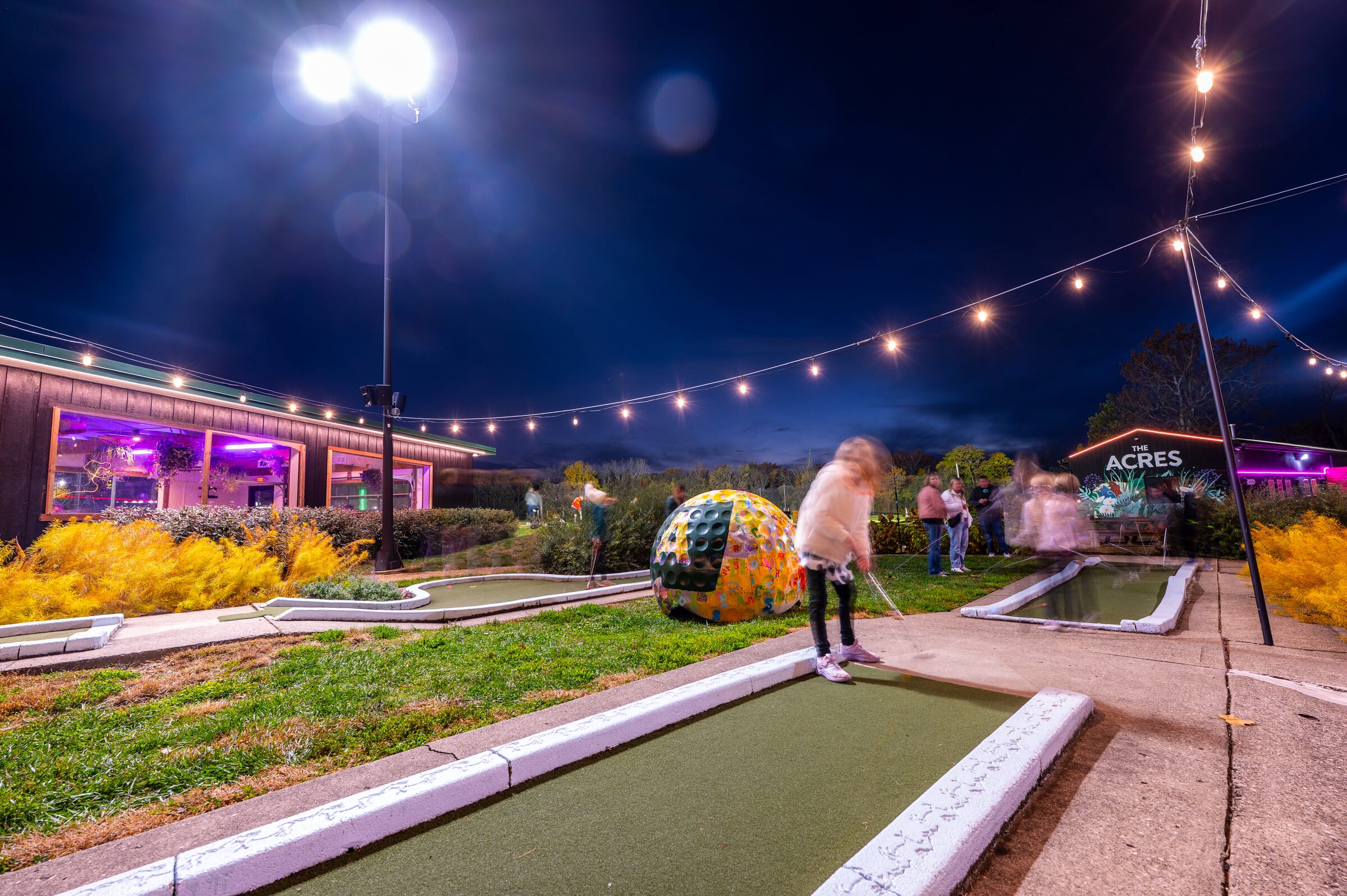 The Acres mini golf course at night under string lights, with a painted ball sculpture and the lit barn